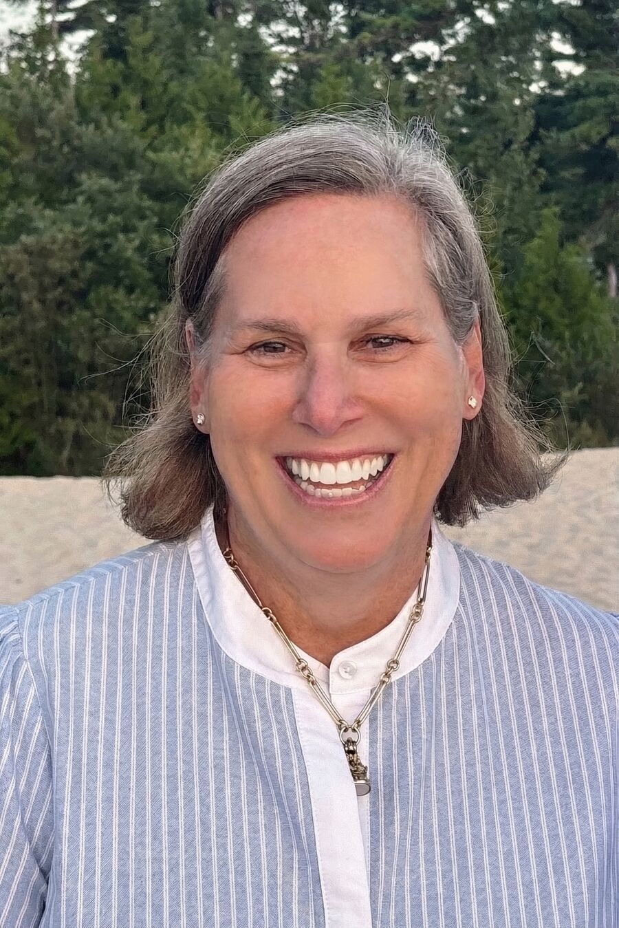 Portrait of Beth Apley Kost, smiling, with evergreens in the background